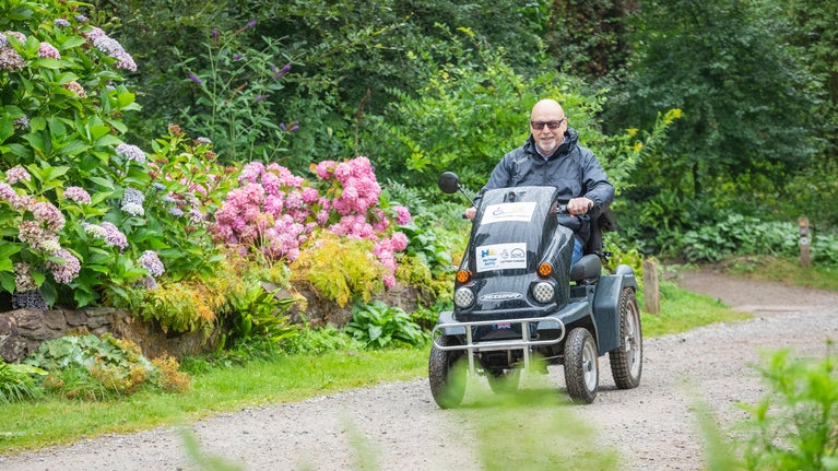 A visitor explores gardens in a tramper, a four wheeled mobility vehicle designed for uneven terrain. Pink and purple flowers and green foliage frame the image.